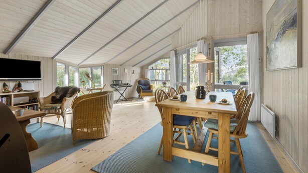 Een ruime kamer met een hoge plafond, een eethoek met een houten tafel en stoelen, een televisie die aan de muur is bevestigd, en grote ramen die natuurlijk licht binnenlaten.