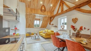 Ein modernes Interieur mit einem gelben Sofa, orangefarbenen Sesseln, einem Esstisch und einer Küchenzeile mit Obstschale.