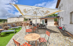 An outdoor patio with stone flooring, wooden tables, and chairs, a large umbrella, and a stone wall.