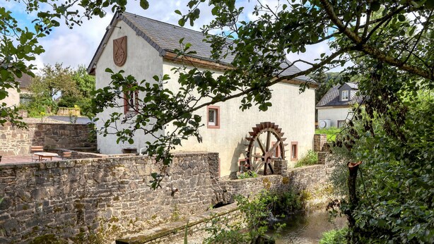 Ein historisches Gebäude mit einem großen Wasserrad, Steinmauern und einer kleinen Brücke über einen Bach.