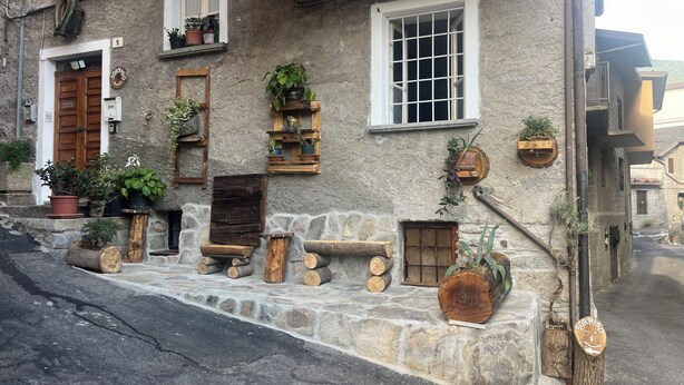 A rustic stone building with wooden benches, potted plants, and a wooden door. - SMILE          (Berzo)