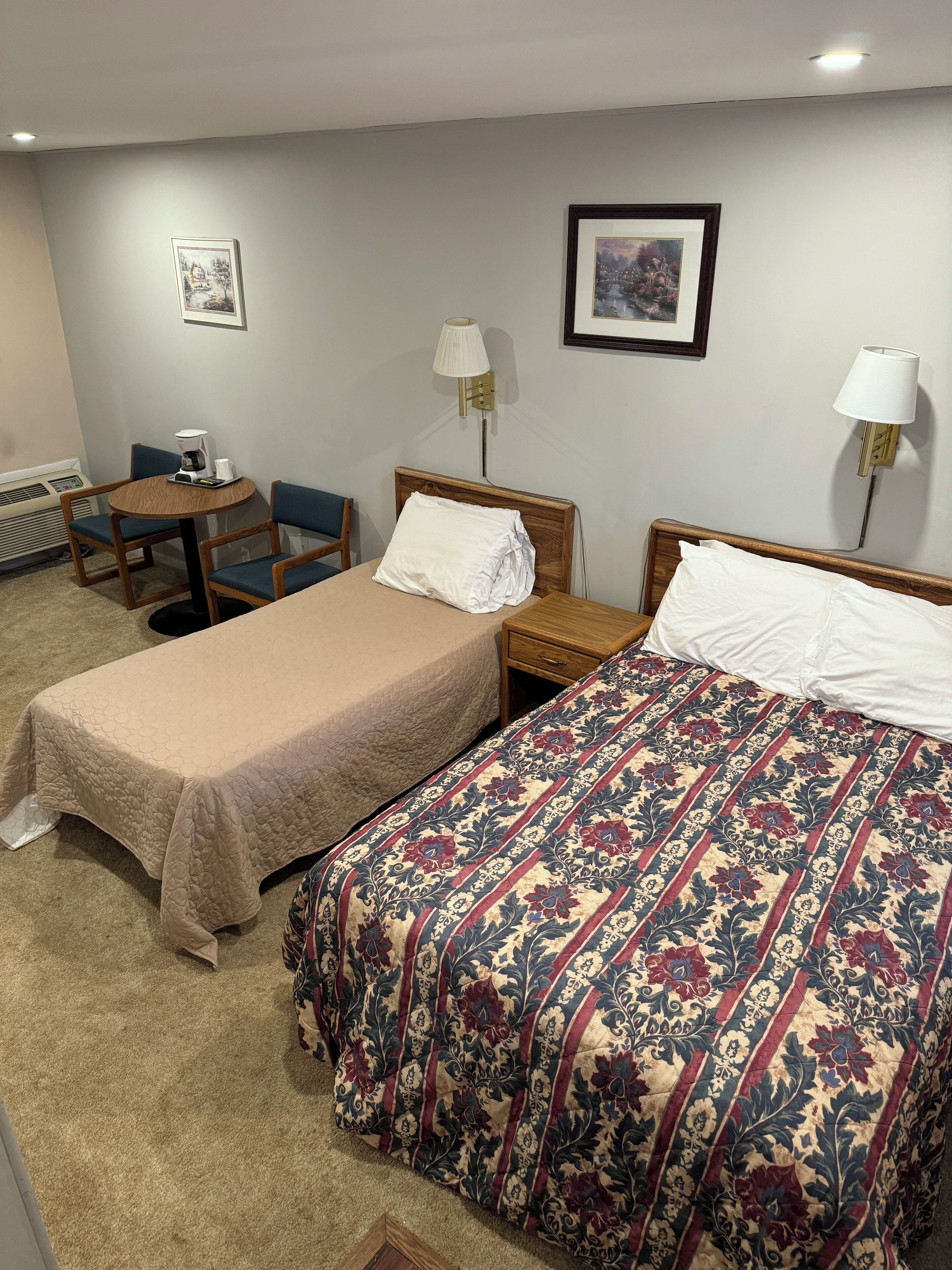 A hotel room with two beds, a small table with a chair, and two wall-mounted lamps.