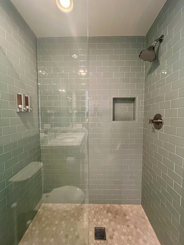 Douche met glazen wand, betegelde muur, douchekop en een klein schap.