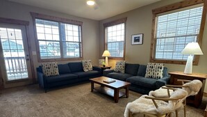 Ein Wohnzimmer mit einem dunkelblauen Sofa, einem Holztisch und einem flauschigen Sessel. Zwei Fenster mit Jalousien, ein Deckenventilator und eine Lampe auf einem Nachttisch aus Holz sind vorhanden.