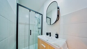 Une salle de bain moderne avec une douche à l’italienne en verre, un meuble-lavabo en bois avec un lavabo blanc et un miroir rond.