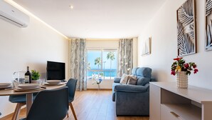 Ein modernes Wohnzimmer mit einer Couch, einem Essbereich mit Tisch und Stühlen, einem Fernseher und Blick aufs Meer.