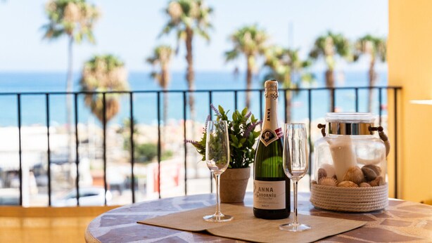 Une table avec une bouteille de vin Anna Codorniu Brut, deux verres et un bocal de noix, sur un balcon donnant sur une plage bordée de palmiers.
