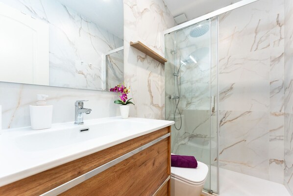 A modern bathroom with a marble backsplash, a wooden vanity, a glass shower enclosure, and a toilet.