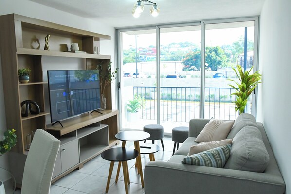 Una sala de estar moderna con un televisor de pantalla plana, un sofá, dos mesas redondas y un balcón con vistas a un estacionamiento y zonas verdes.