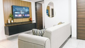 A modern living area with a flat-screen TV mounted on a wooden panel, a sofa with patterned cushions, a mirror, and a door leading to another room. - Modern & Spacious APT - Wifi, Gym, Pool, A/C (San Salvador)