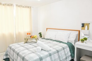 Habitación de hotel ordenada con una cama, una mesita de noche con fruteros, una toalla enrollada y una pequeña planta en maceta.
