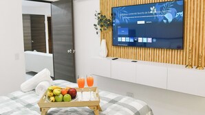 A modern hotel room with a flat-screen TV mounted on a wooden panel wall, a bed with a checkered bedspread, a tray with fruits and a glass of orange juice, and a white bedside table with a vase of green leaves. - Modern & Spacious APT - Wifi, Gym, Pool, A/C (San Salvador)