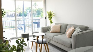 A modern living room with a grey sofa, two small round tables, and a large sliding glass door leading to an outdoor balcony. - Modern & Spacious APT - Wifi, Gym, Pool, A/C (San Salvador)