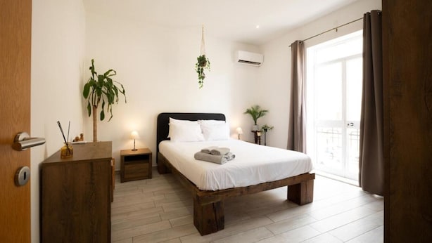 A bedroom with a wooden bed, bedside tables, a hanging plant, an air conditioning unit, and a view through a glass door.