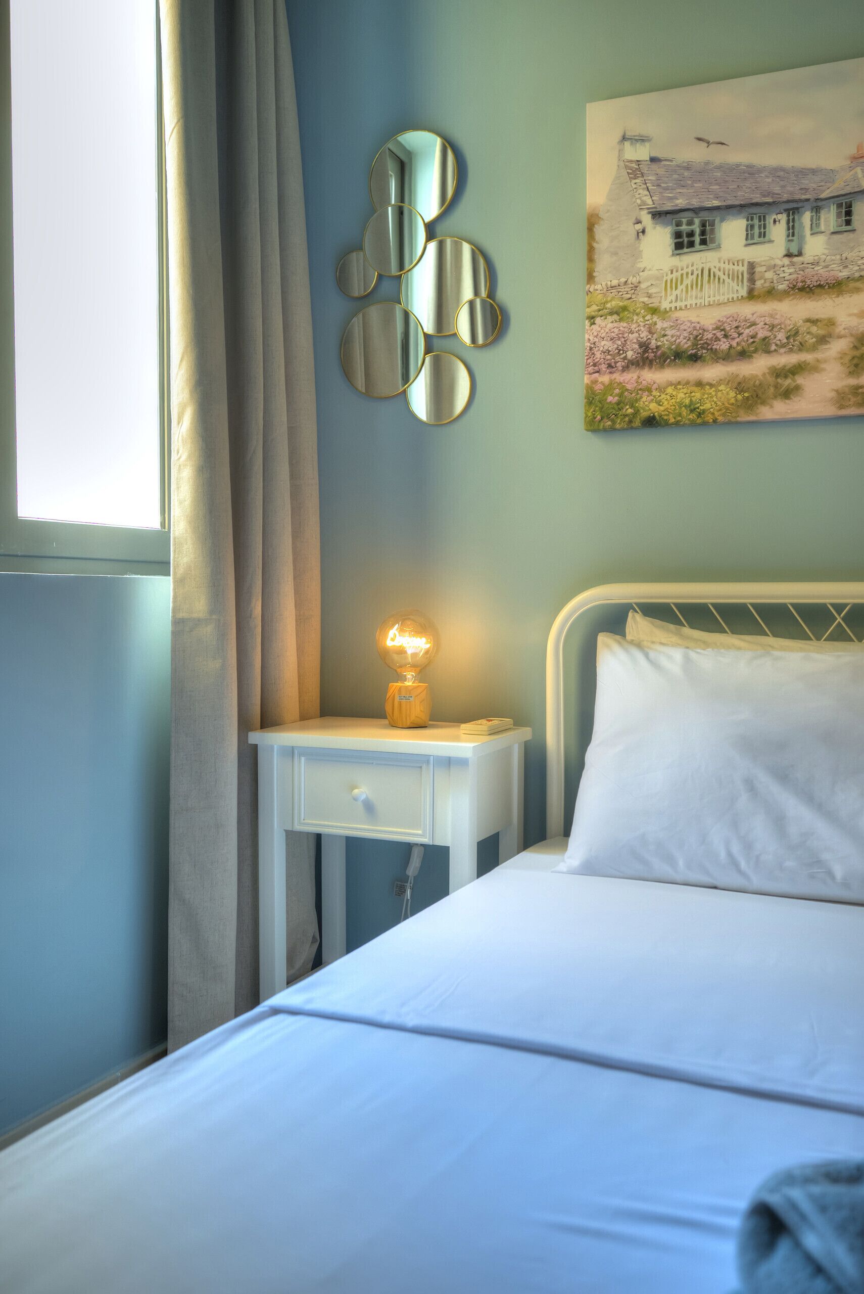 A bedroom with a bed, a bedside table with a lamp, a wall-mounted mirror, and a painting of a house.