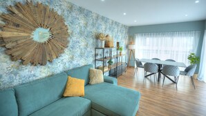 A modern living room with a teal sofa, a large wooden wall art piece, a glass cabinet with plants, and a dining area with a table and chairs. - Stylish, large 3bedroom, seafront terrace GOSLM-13 (Sliema)