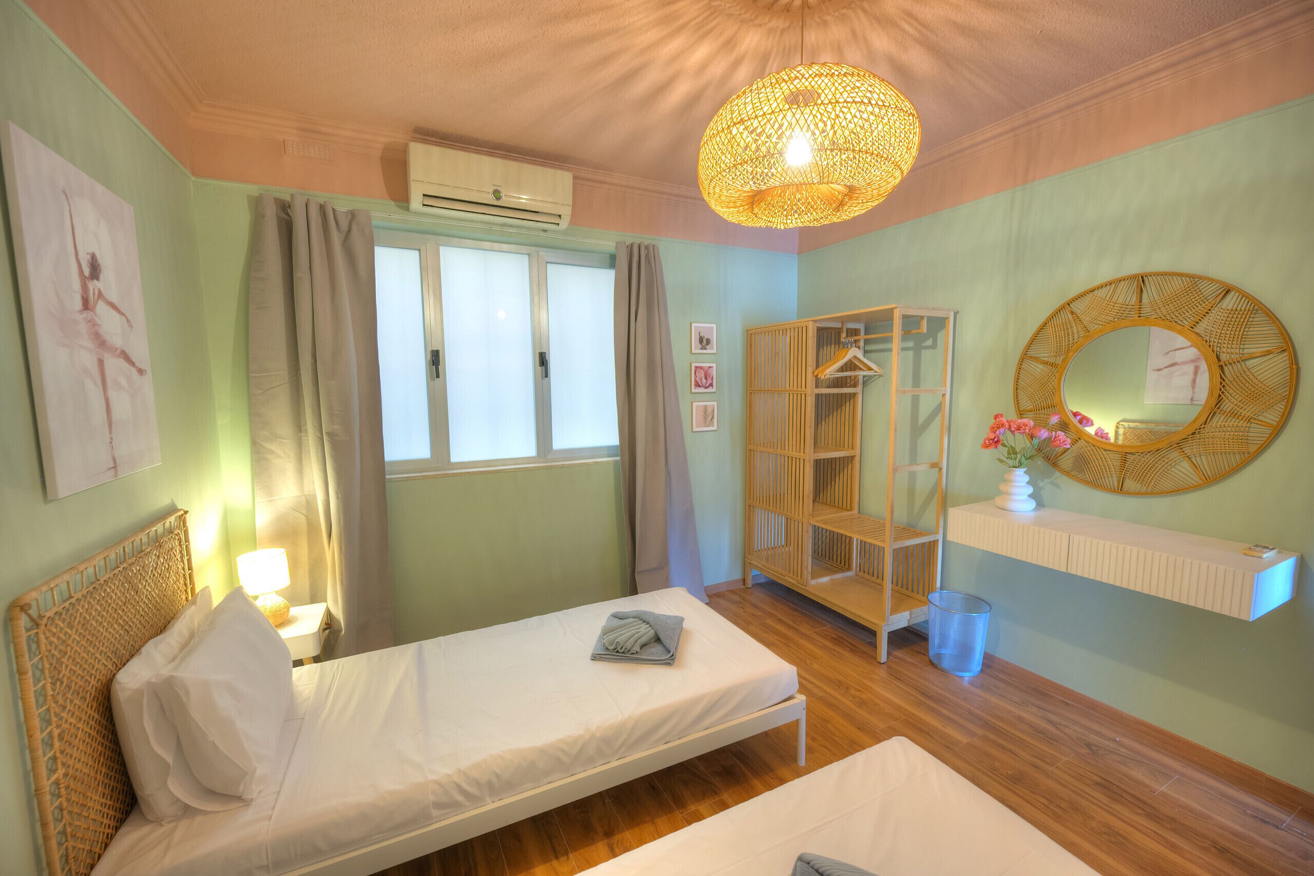 A room with two beds, a wardrobe, a mirror, a hanging light fixture, and a window with curtains.