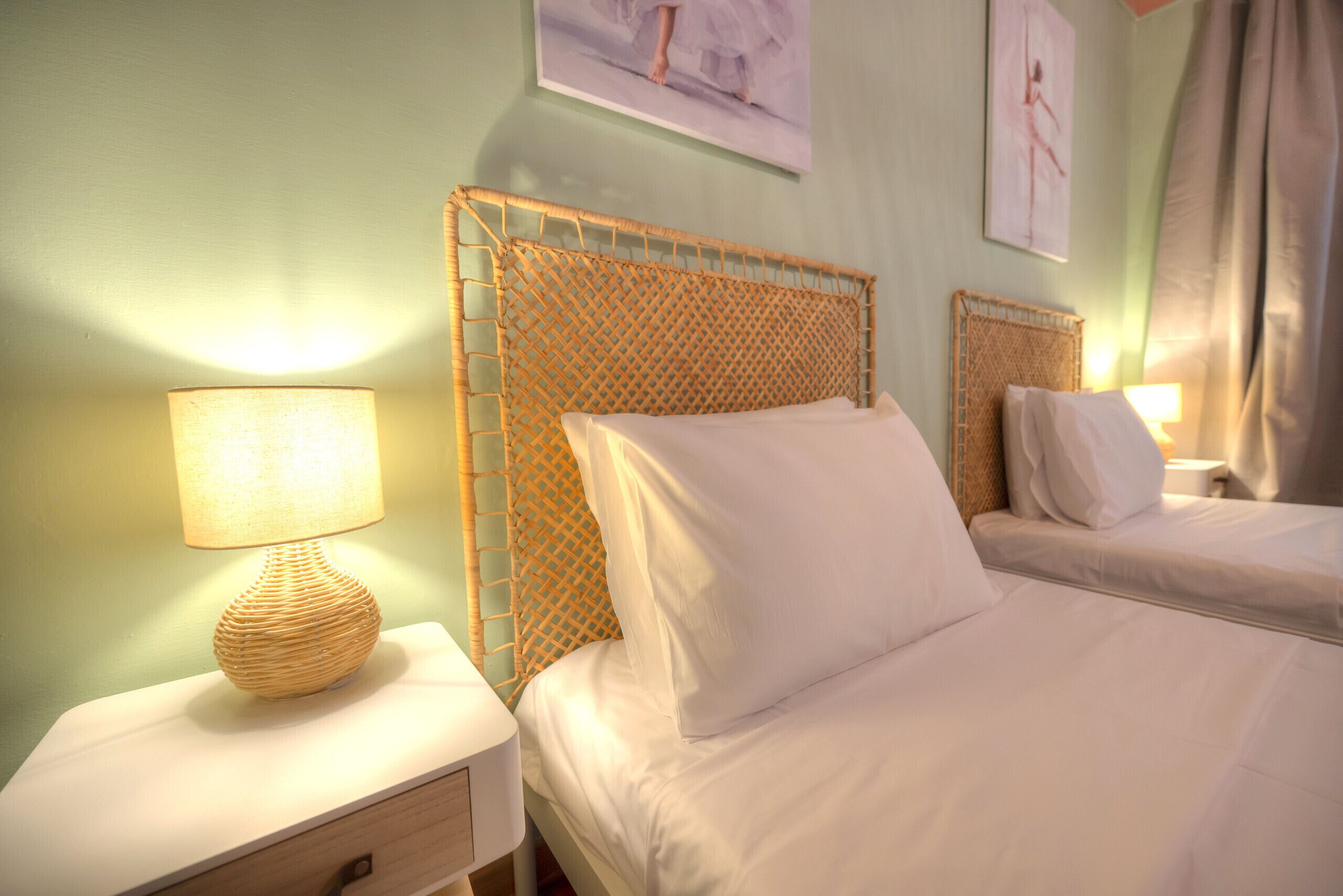 Habitación de hotel con dos camas, una mesita de noche de mimbre con una lámpara y obras de arte en las paredes.