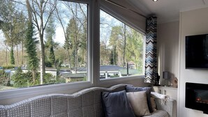 A modern living room with a patterned sofa, a fireplace, and a large window offering a view of trees and houses. - New luxury chalet at 't Veluws Hof AC/kids++/wifi (Hoenderloo)