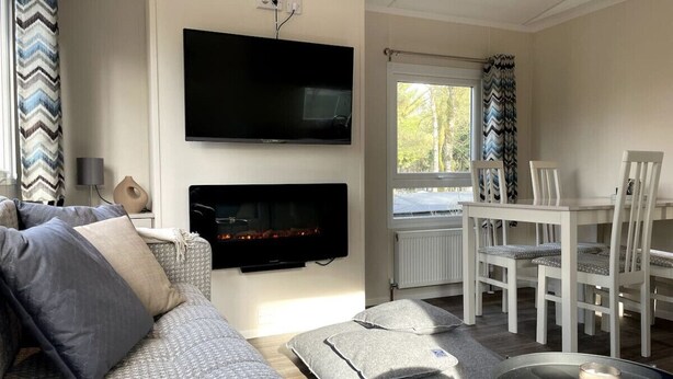 A living room with a sofa, a flat-screen TV mounted on the wall, a fireplace, and a dining area with white chairs and a table. - New luxury chalet at 't Veluws Hof AC/kids++/wifi (Hoenderloo)
