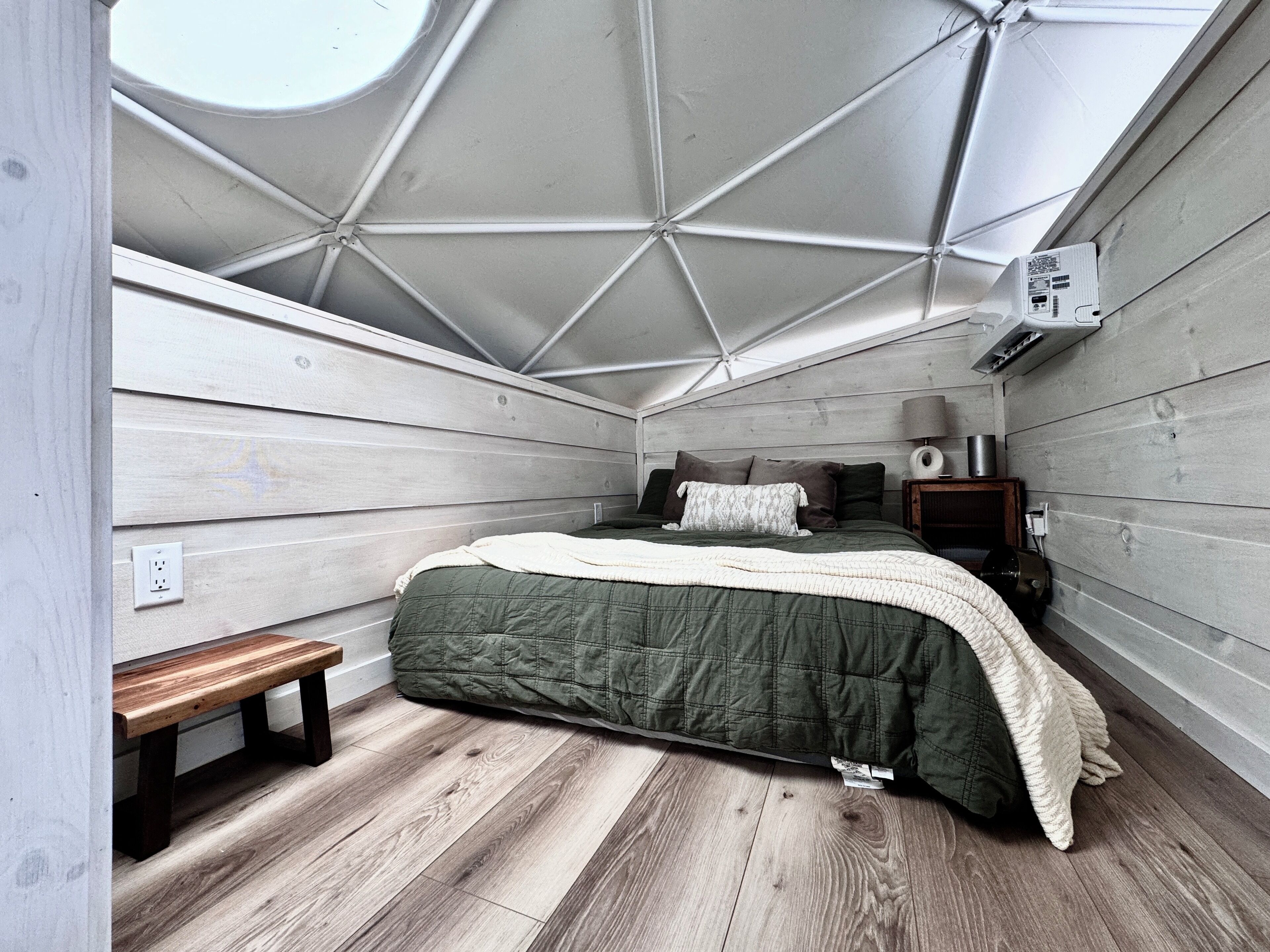 Un dormitorio con techo inclinado, una cama con colcha verde, un banco de madera, una mesita de noche con lámpara y un aire acondicionado instalado en la pared.