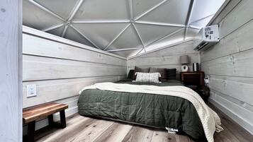 Un dormitorio con techo inclinado, una cama con colcha verde, un banco de madera, una mesita de noche con lámpara y un aire acondicionado instalado en la pared.