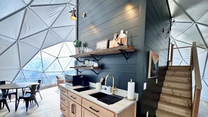 Een moderne keuken met een centraal eiland, houten kastjes en een ingebouwde gootsteen. Boven de gootsteen zijn planken en rechts een trap.