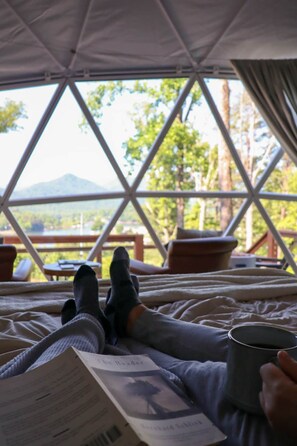 Une personne allongée sur un lit à l’intérieur d’une structure ressemblant à une tente, avec un livre et une tasse à portée de main, et qui a devant elle une vue imprenable sur un paysage extérieur avec des arbres et une montagne.