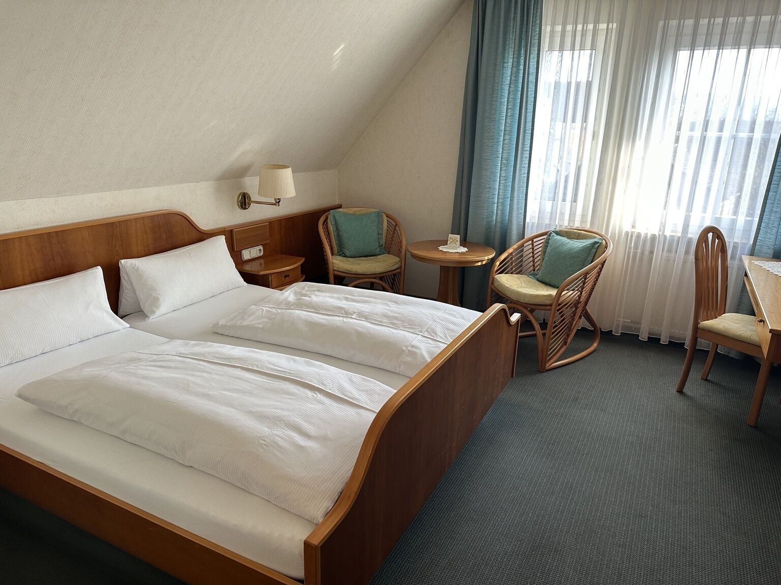 A hotel room with a large bed, two bedside tables, a lamp, two wicker chairs, a small round table, and a view of the outside through sheer curtains.