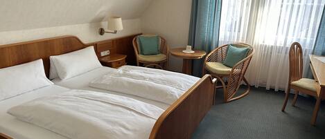 Habitación de hotel con una cama grande, dos mesitas de noche, una lámpara, dos sillas de mimbre, una mesita redonda y vistas al exterior a través de cortinas traslúcidas.