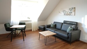 A modern living room with a dark leather sofa, a wooden coffee table, and two green armchairs. A white radiator and a window with curtains are visible in the background. - G. Hotel Müller (Lichtenfels)