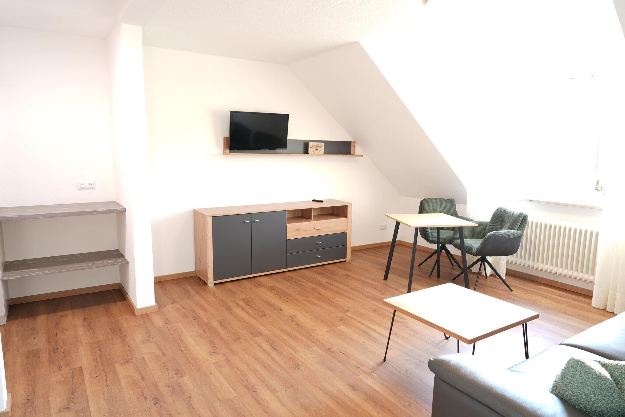 Een moderne woonkamer met een houten vloer, een grijze bank, een kleine tafel en een aan de muur bevestigde televisie.