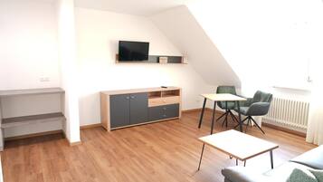 Een moderne woonkamer met een houten vloer, een grijze bank, een kleine tafel en een aan de muur bevestigde televisie.