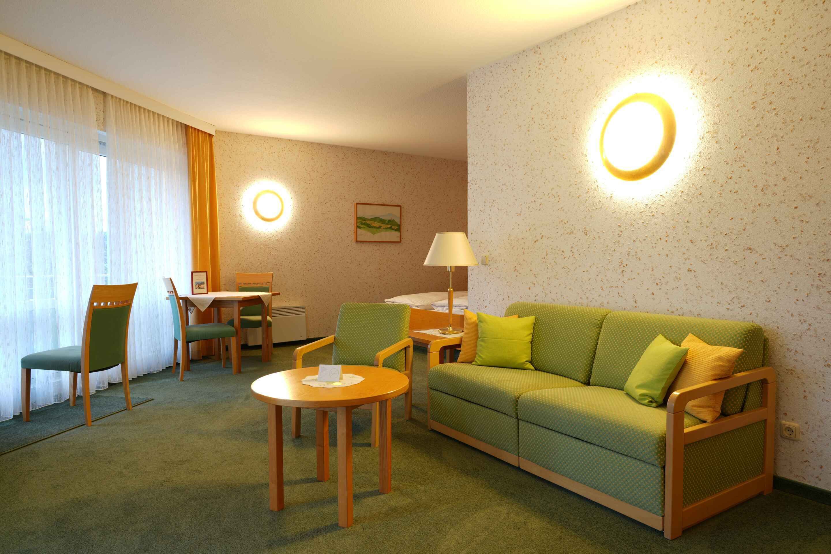 A hotel room with a green sofa, two chairs, a small table, a lamp, and a painting on the wall.