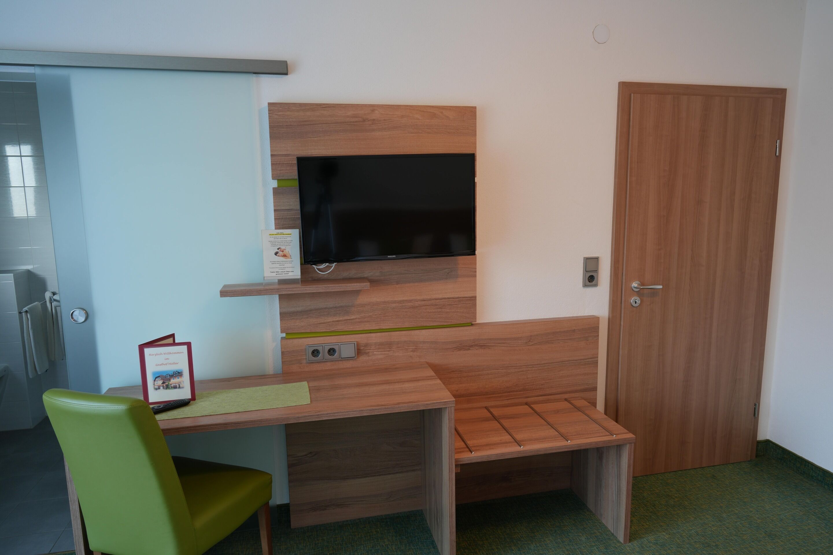 Una habitación pequeña con un escritorio de madera, una silla verde, un televisor montado en la pared y una puerta de madera.
