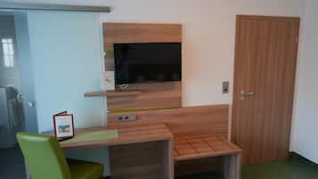 Una habitación pequeña con un escritorio de madera, una silla verde, un televisor montado en la pared y una puerta de madera.