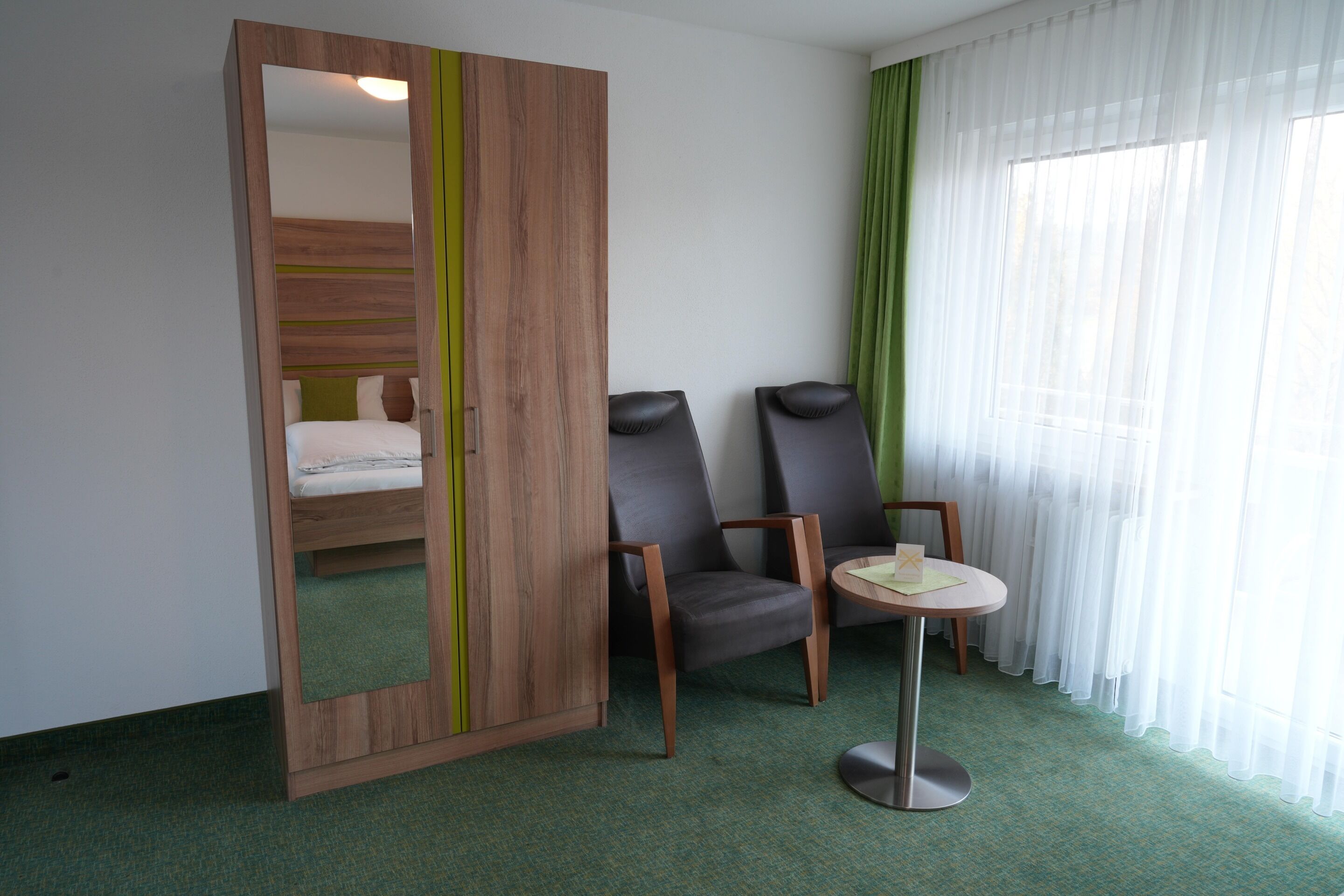 Une chambre d’hôtel avec un lit, un miroir, deux chaises, une petite table et une fenêtre avec des rideaux légers.