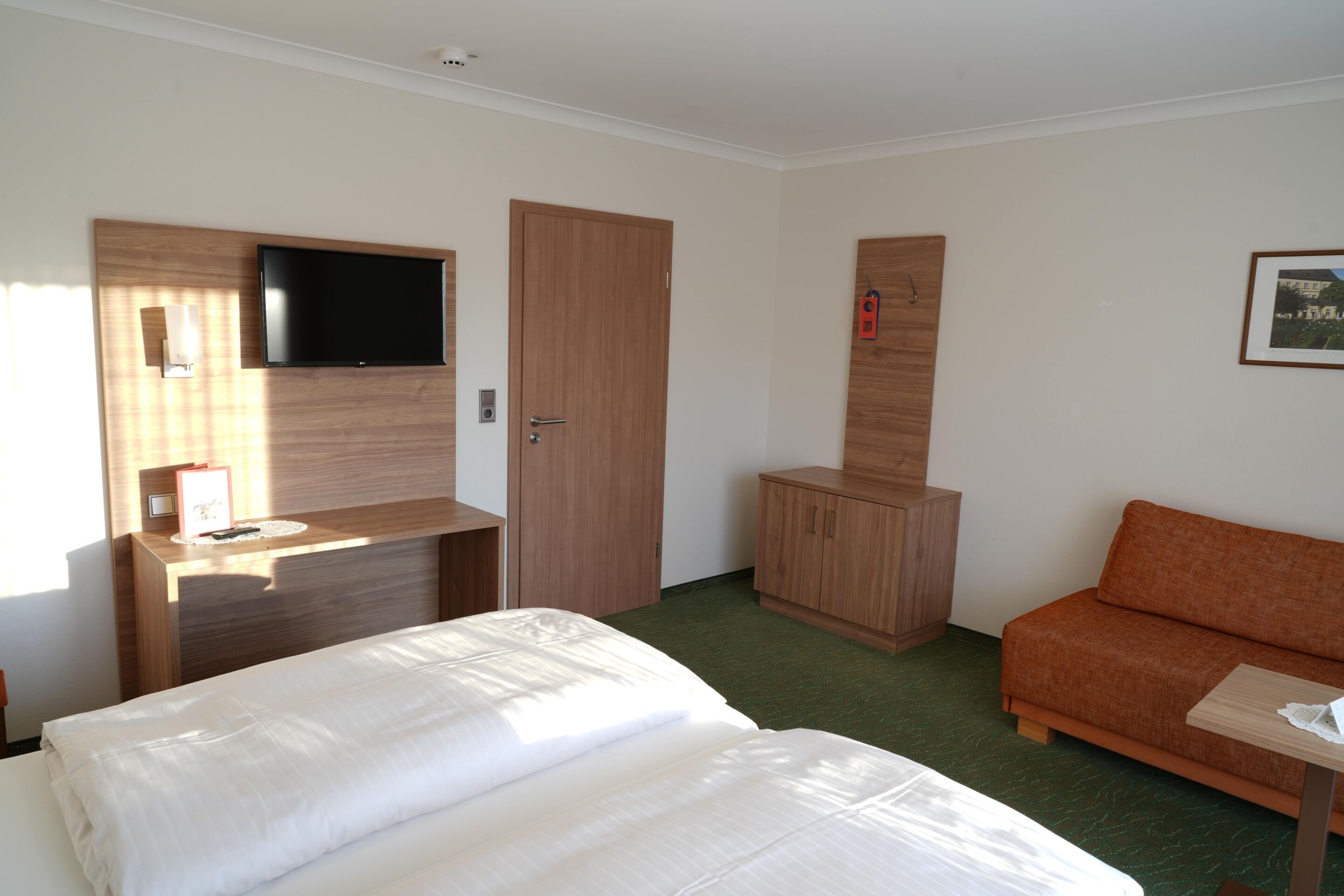 A hotel room with a bed, a TV mounted on a wooden panel, a wooden wardrobe, and a small table with a lamp.