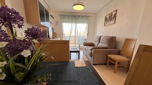A living room with a sofa, a chair, a small table, and a TV. There is a window with curtains, a plant with purple flowers, and a rug on the floor. - ACV - Costa Azahar II-2nd floor line 6 north 2 (OROPESA)