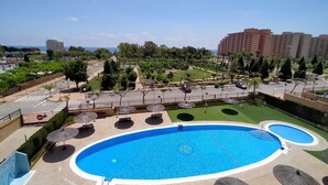 A swimming pool with a circular design, surrounded by lounge chairs and umbrellas, set in a landscaped area with trees and a view of a cityscape. - ACV - Costa Azahar II-2nd floor line 6 north 2 (OROPESA)