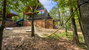 A wooden cabin with a slide, surrounded by trees and a hammock.