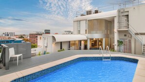 Una piscina sul tetto con vista sulla città, una zona relax all'aperto coperta e un edificio moderno con facciata in vetro.