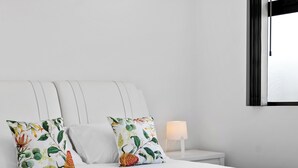 Ein ordentlich bezogenes Bett mit weißen Leinentextilien, zwei blumigen Kissen, einer türkisfarbenen Tagesdecke und einem weißen Nachttisch mit einer Lampe.