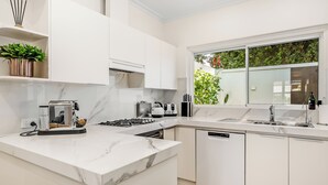 A modern kitchen with white cabinetry, a marble countertop, a built-in dishwasher, and a window with a view of greenery.