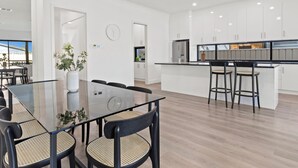 A modern kitchen with white cabinets, a central island with bar stools, and a dining area with a glass table and chairs. - Swainson on Lexington (Henley Beach South)
