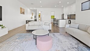A modern living room with a sofa, ottoman, and coffee table. There is a dining area with a table and chairs, and a kitchen in the background. - Swainson on Lexington (Henley Beach South)