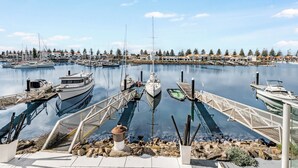 Uma marina com barcos atracados, céu claro e fileira de casas ao fundo.