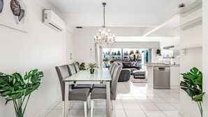 Uma cozinha moderna com máquina de lavar louça, uma área de jantar com mesa de vidro e cadeiras cinzas, e vista para o exterior.