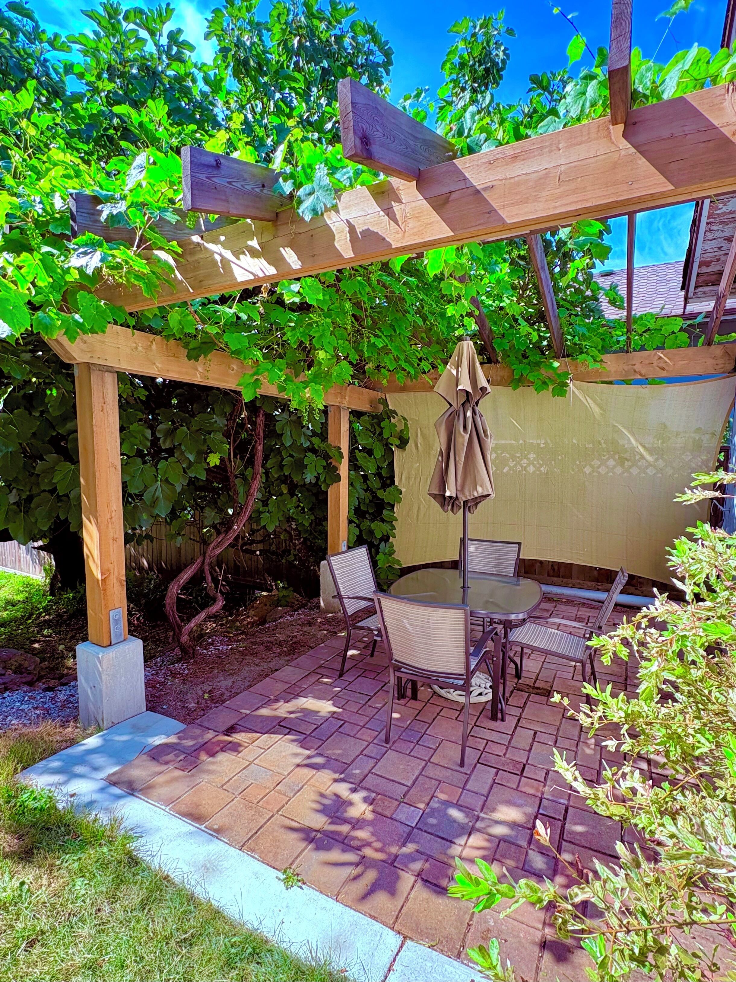 A wooden pergola with a table and chairs underneath, surrounded by greenery.