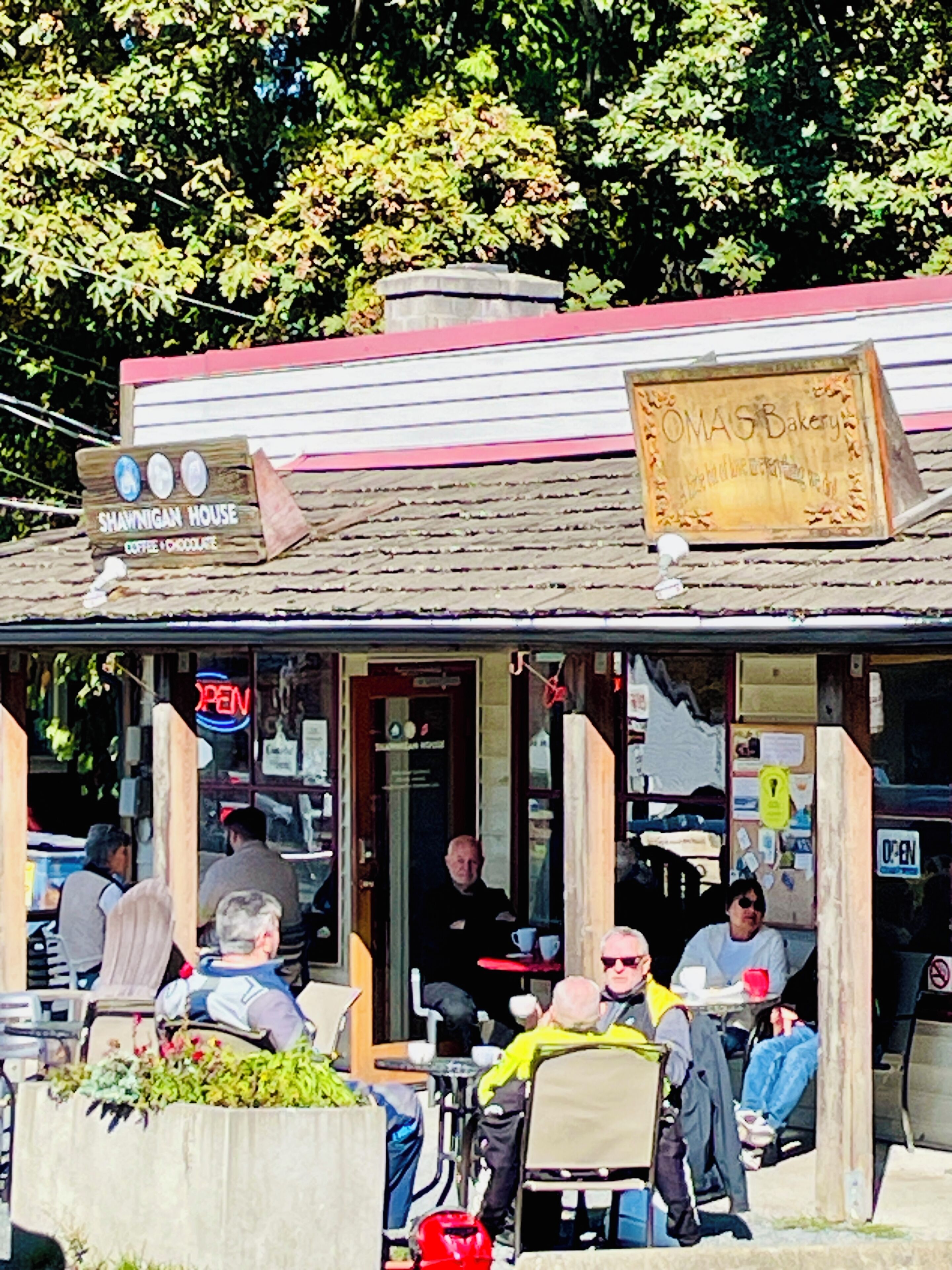 Un pequeño café al aire libre con fachada de madera rústica, un cartel que dice 'OMAS Bakery' y un letrero de neón que indica 'ABIERTO'.